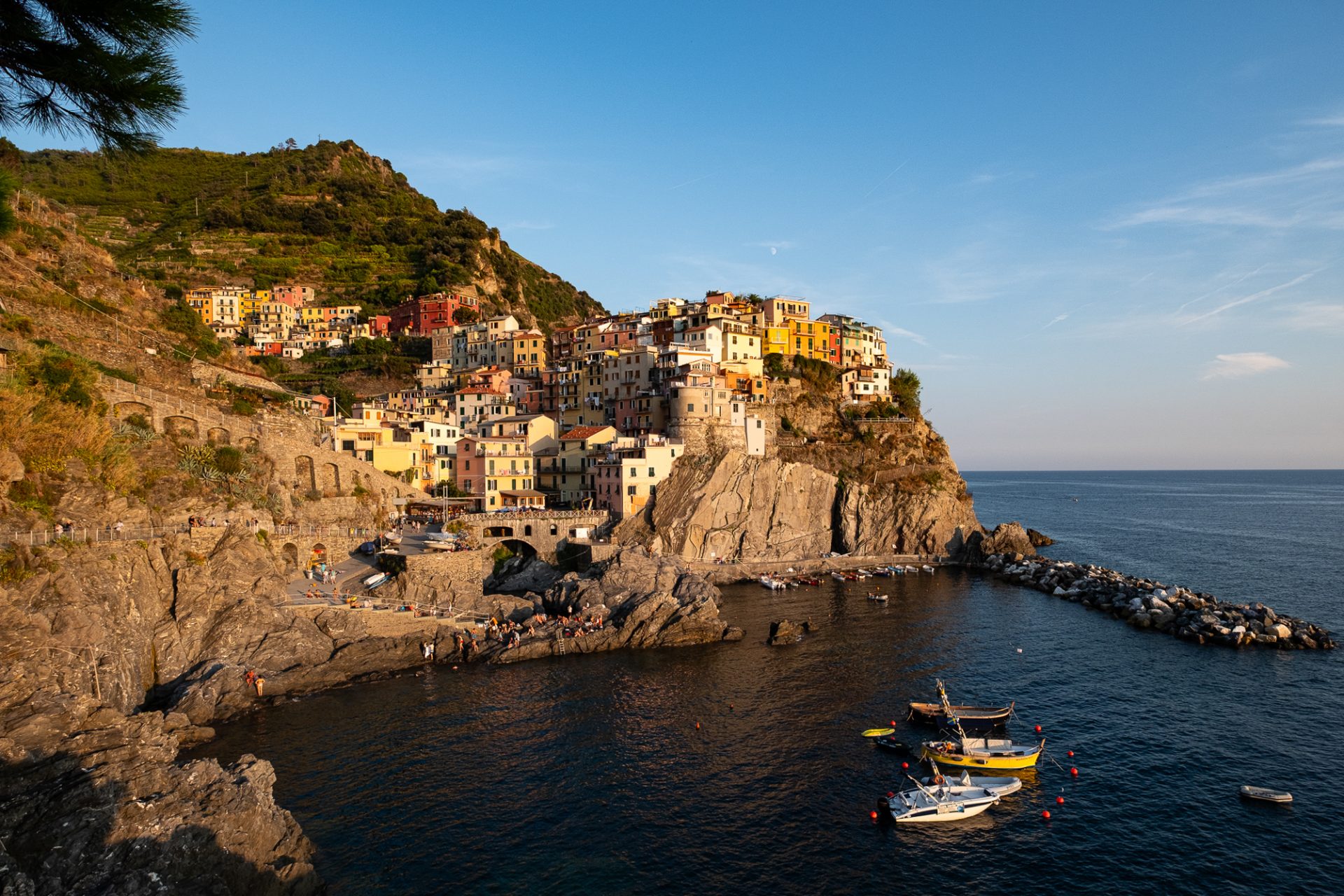 Color photo of Manarola's pastel houses stacked on a cliff above the harbor at golden hour, boats in the cove, Cinque Terre.