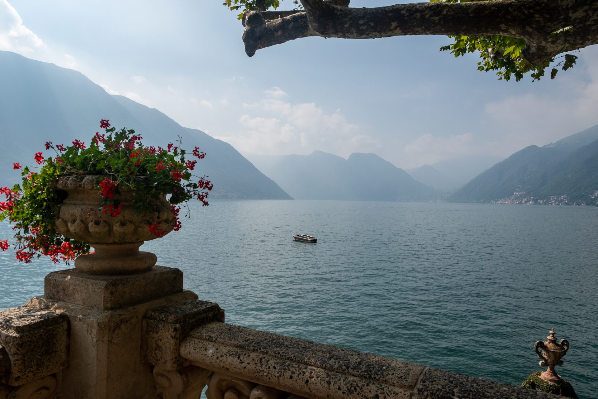 Color photo of a stone balustrade urn spilling red geraniums above Lake Como, hazy mountains and a single boat beyond.