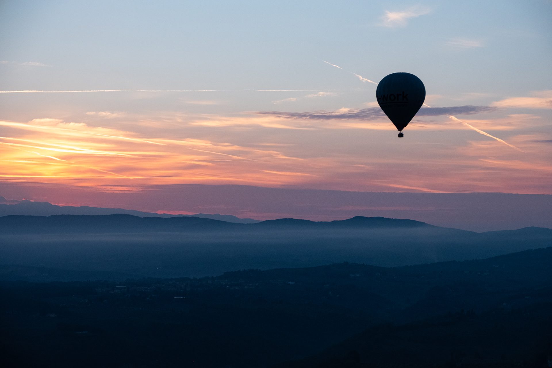 A hot air balloon silhouetted against a pink dawn sky above misted Tuscan hills