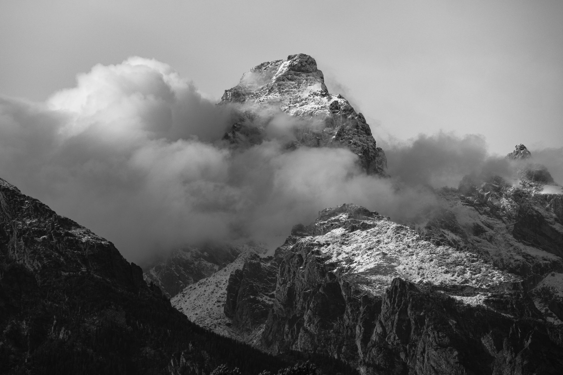 Black and white photo of a snow-covered Grand Teton peak breaking through storm clouds, lit along the summit.