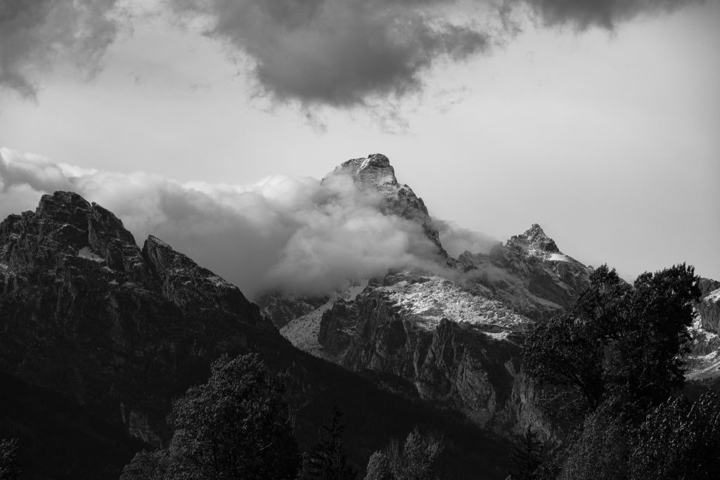 Black and white photo of a Grand Teton peak wrapped in low clouds with a dark forested ridge in the foreground.