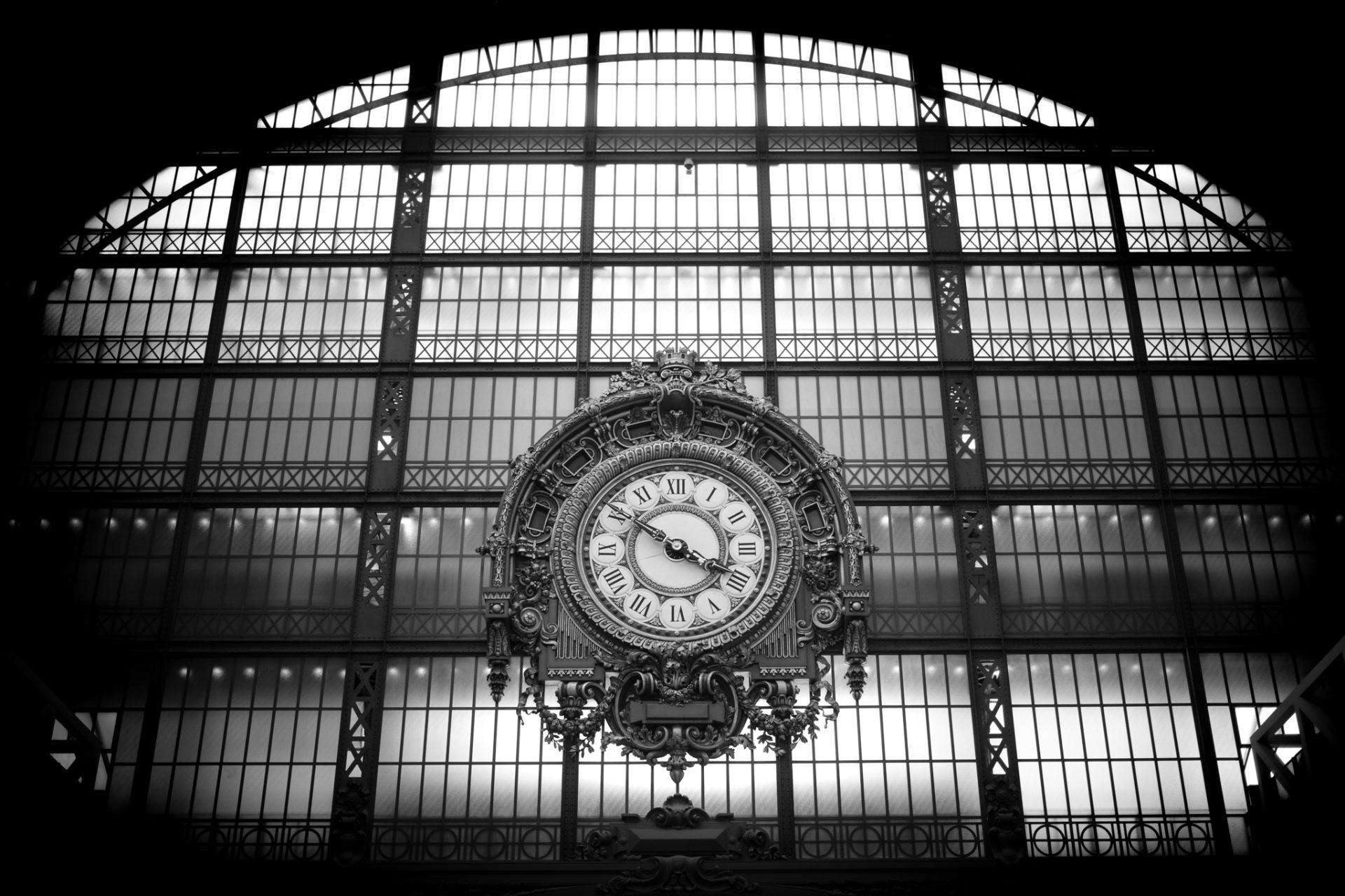 Black and white photograph of the ornate station clock inside the Musée d'Orsay, set against the great arched glass wall of the former railway hall.