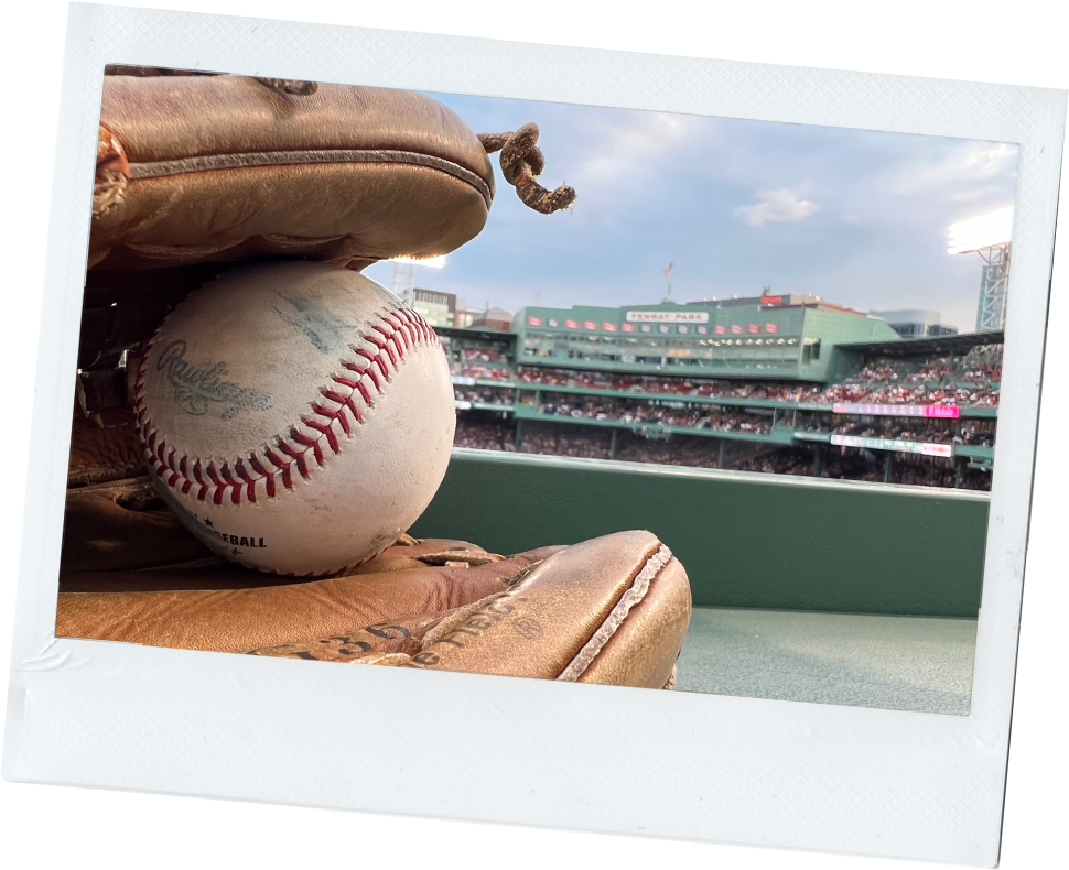 A baseball sitting in a worn leather glove with Fenway Park in the background.