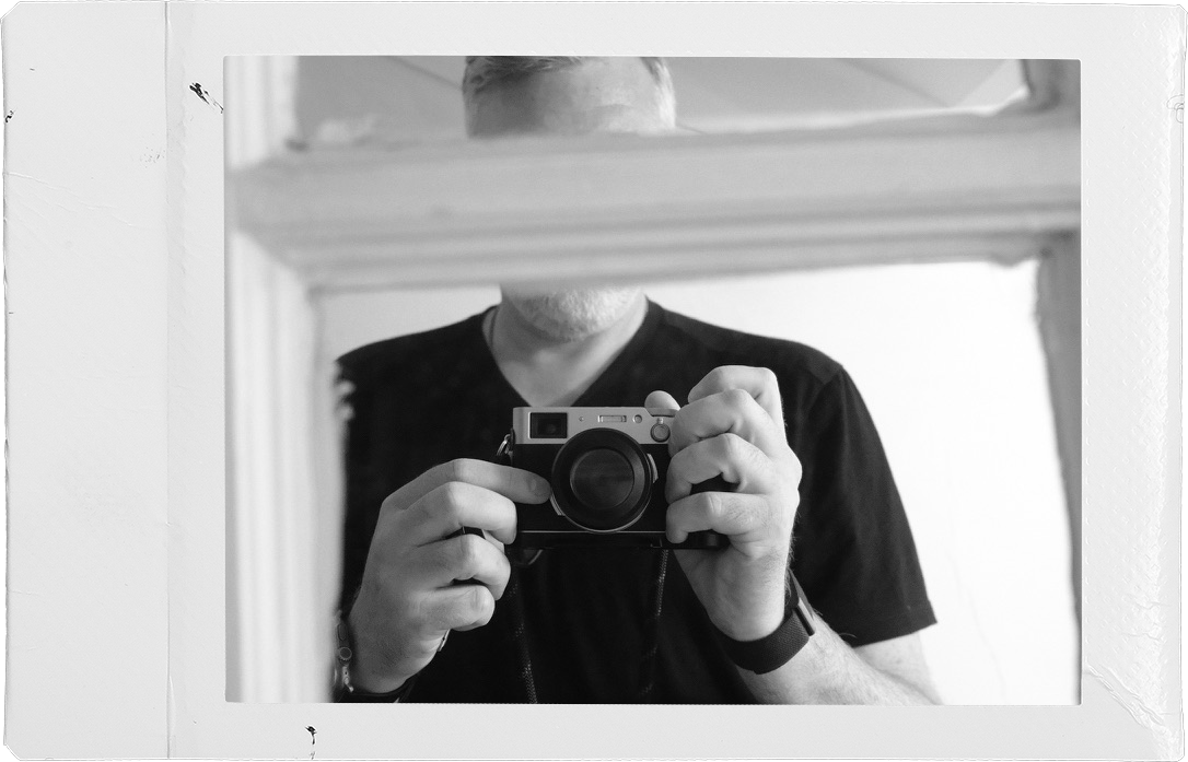 Black and white mirror self-portrait of a man holding a camera up to his face, framed by a white instant-film border.